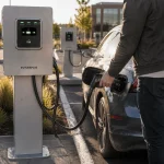Person inserting a CCS charging gun into an electric sedan in a parking lot at sunset, with a charging station on the left